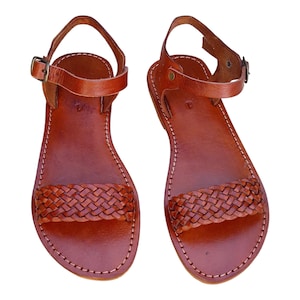 Peut inclure: Paire de sandales en cuir marron avec une bride tressée sur les orteils et une bride de cheville avec une boucle. Les sandales ont une semelle cousue et sont sur fond blanc.