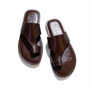 May include: A pair of brown leather sandals with a thong-style toe post and a wide strap across the top of the foot. The sandals have a stitched sole and a textured footbed. The sandals are displayed on a white background.