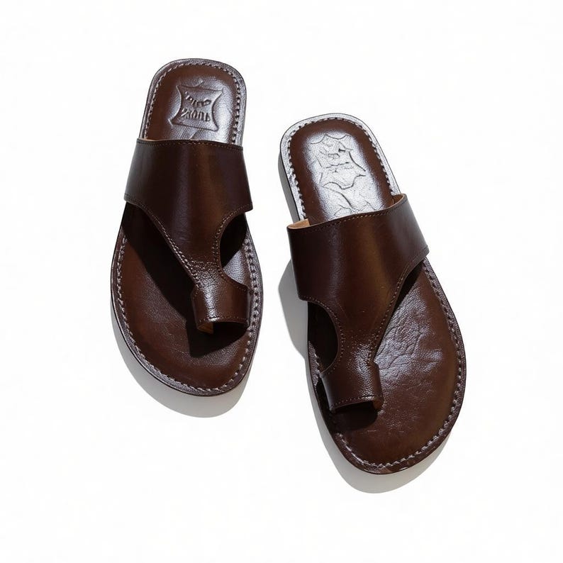 May include: A pair of brown leather sandals with a thong-style toe post and a wide strap across the top of the foot. The sandals have a stitched sole and a textured footbed. The sandals are displayed on a white background.