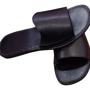 May include: A pair of black leather slide sandals. The sandals have a simple design with a wide strap across the top and a stitched sole. The sandals are approximately 10 inches long.