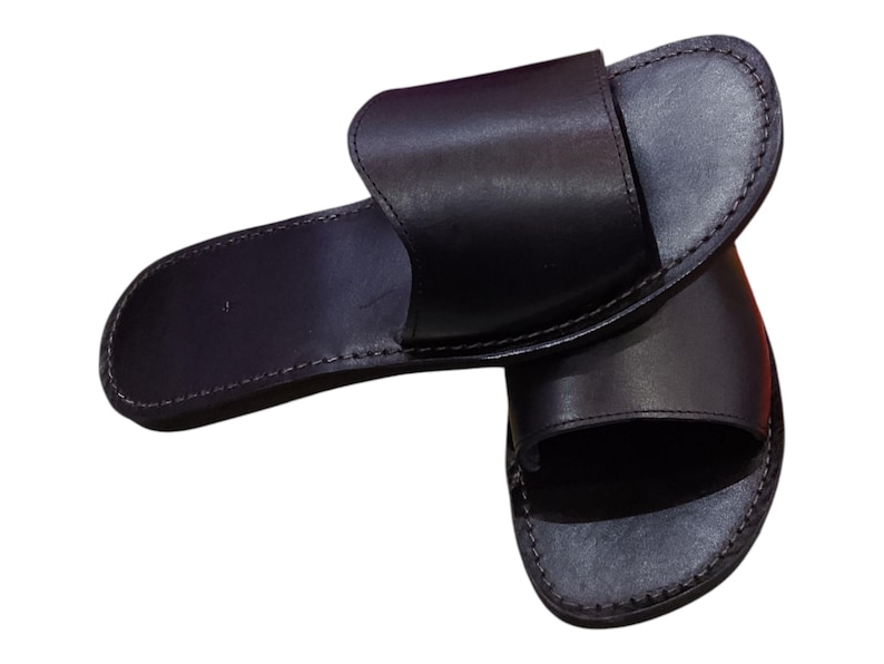 May include: A pair of black leather slide sandals. The sandals have a simple design with a wide strap across the top and a stitched sole. The sandals are approximately 10 inches long.