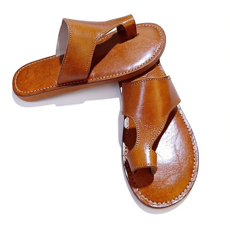 May include: A pair of brown leather sandals with a toe loop design. The sandals have a stitched sole and a strap that goes over the top of the foot. The leather has a glossy finish.