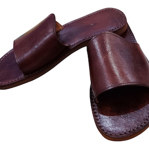 May include: A pair of brown leather slide sandals. The sandals have a simple design with a wide strap across the top of the foot and a stitched sole. The leather has a slightly textured appearance.