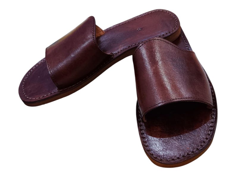 May include: A pair of brown leather slide sandals. The sandals have a simple design with a wide strap across the top of the foot and a stitched sole. The leather has a slightly textured appearance.