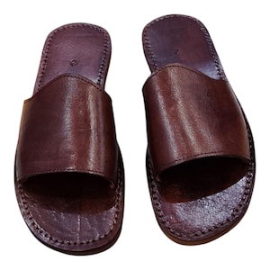 May include: A pair of brown leather sandals. The sandals have a slip-on design with a wide strap across the top of the foot. The leather has a textured appearance and is stitched around the edges. The sandals are designed for casual wear.