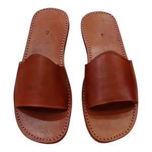 May include: A pair of brown leather slide sandals. The sandals have a simple design with a wide strap across the top of the foot and a stitched sole. The size "9" is visible on the heel. The sandals are new.