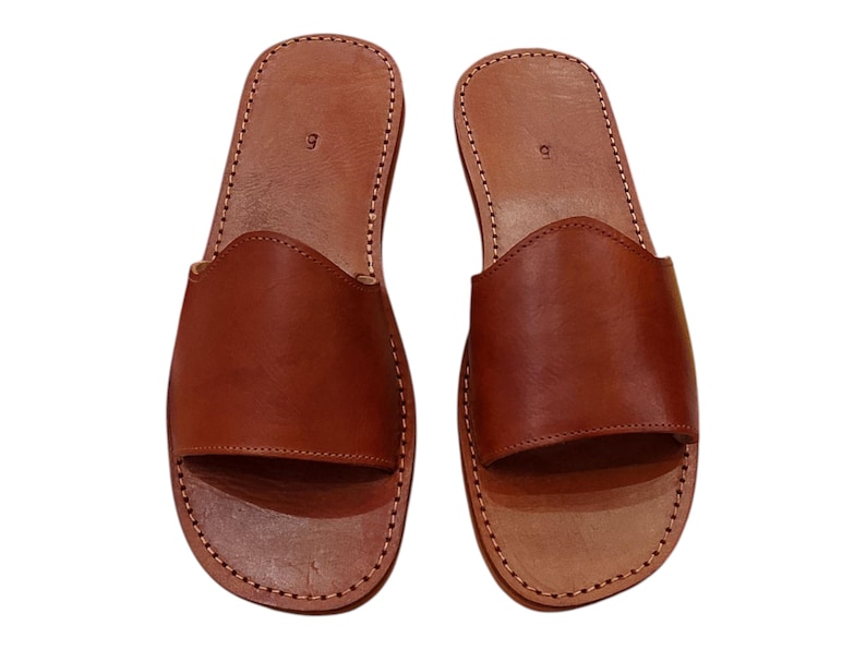 May include: A pair of brown leather slide sandals. The sandals have a simple design with a wide strap across the top of the foot and a stitched sole. The size "9" is visible on the heel. The sandals are new.