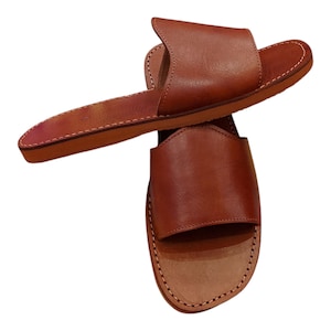 May include: A pair of brown leather sandals. The sandals have a simple design with a strap across the top of the foot and a flat sole. The edges are stitched with white thread. The sandals are made of a smooth, polished leather.