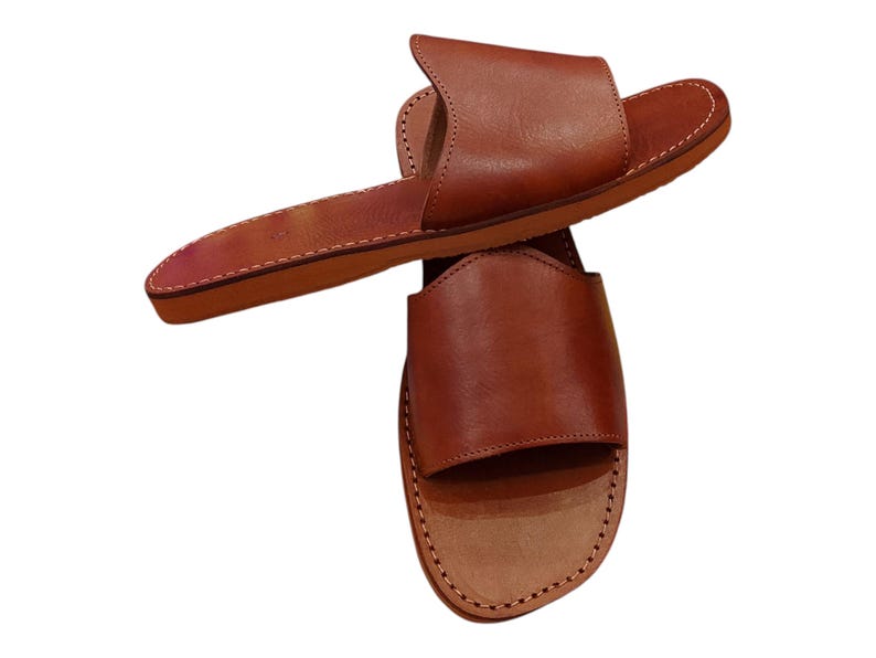May include: A pair of brown leather sandals. The sandals have a simple design with a strap across the top of the foot and a flat sole. The edges are stitched with white thread. The sandals are made of a smooth, polished leather.