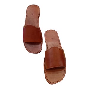 May include: A pair of brown leather slide sandals. The sandals have a simple design with a wide strap across the top of the foot and a stitched sole. The leather is a warm, medium brown color.