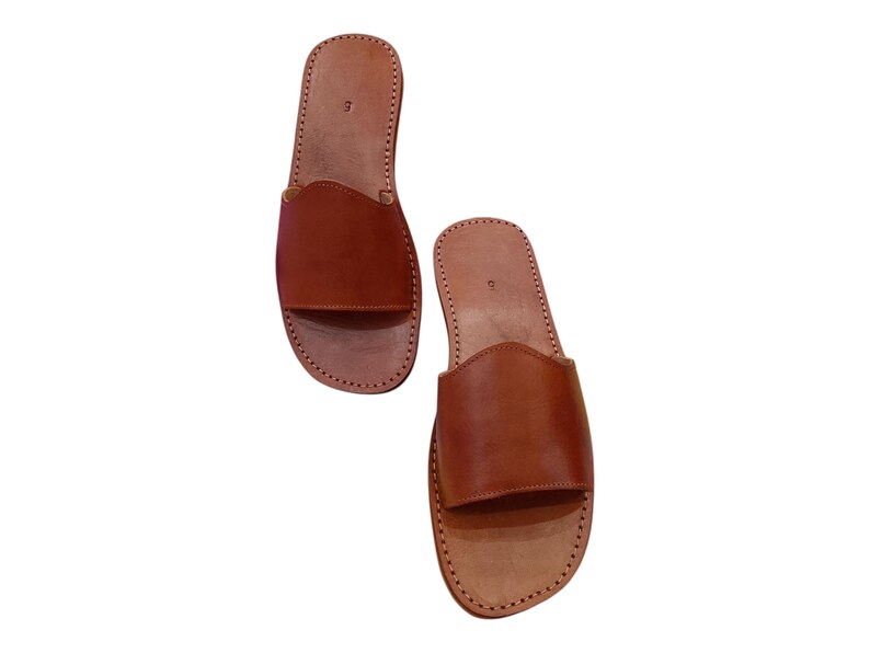 May include: A pair of brown leather slide sandals. The sandals have a simple design with a wide strap across the top of the foot and a stitched sole. The leather is a warm, medium brown color.