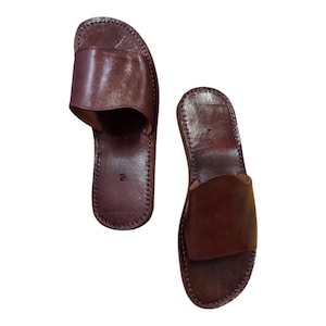 May include: A pair of brown leather slide sandals. The sandals have a simple design with a strap across the top and are stitched around the edges. The sandals are a dark brown color.