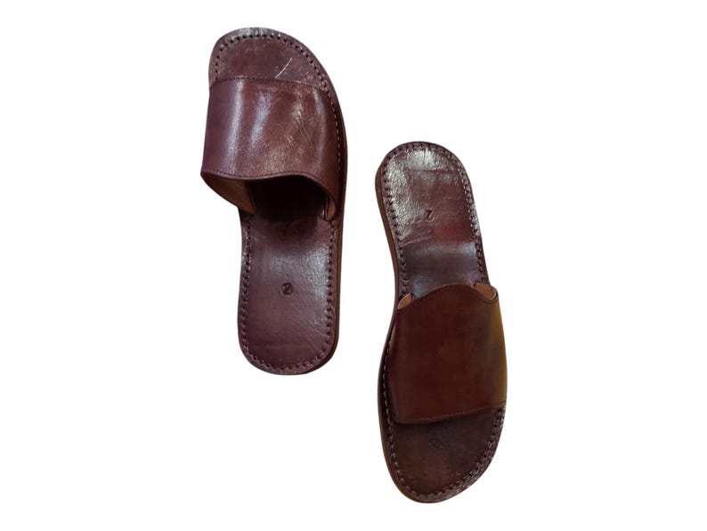 May include: A pair of brown leather slide sandals. The sandals have a simple design with a strap across the top and are stitched around the edges. The sandals are a dark brown color.