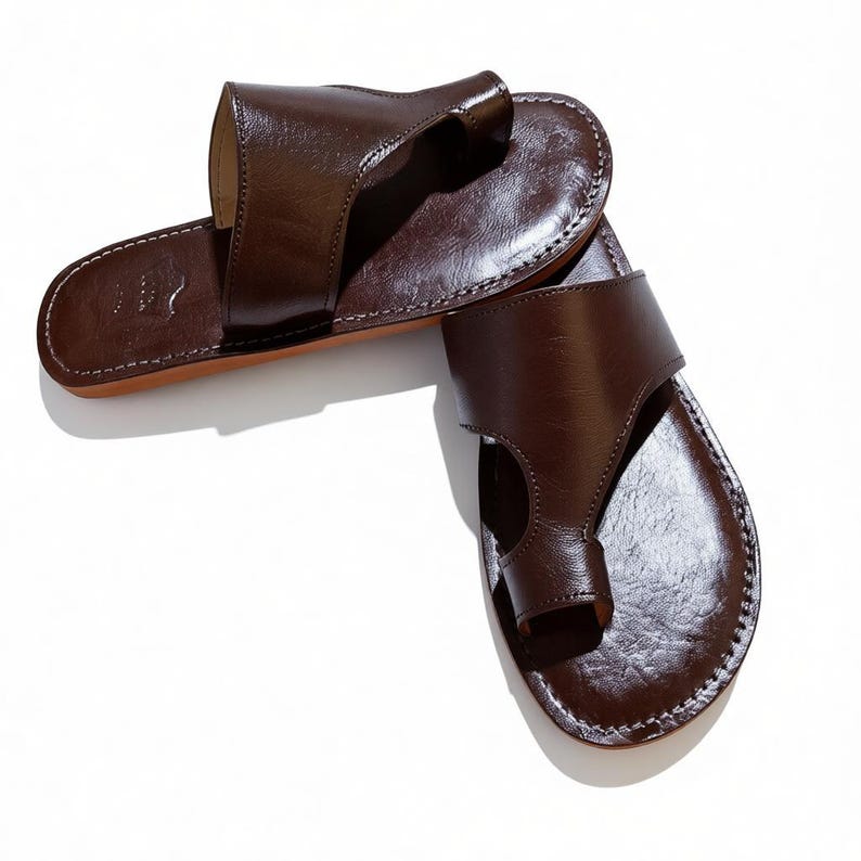 May include: A pair of brown leather sandals with a thong-style toe post and a wide strap across the top of the foot. The sandals have a stitched border and a light brown sole. The leather has a glossy finish.
