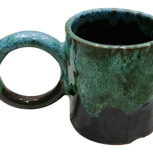 Może przedstawiać: Ręcznie wykonany ceramiczny kubek z dużym, zaokrąglonym uchwytem. Kubek ma gradient kolorów, przechodzący od ciemnoniebieskiego na obrzeżu do zielonego i czarnego u podstawy. Wnętrze jest ciemne.