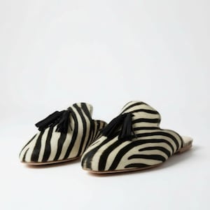 May include: A pair of zebra-striped slip-on shoes with black tassels. The shoes have a rounded toe and a low heel. The zebra pattern is black and white, and the tassels are black.