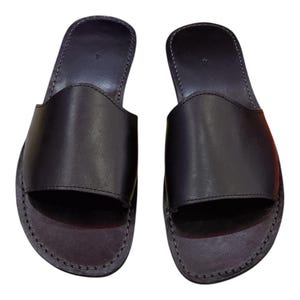 May include: A pair of black leather slide sandals. The sandals have a simple design with a wide strap across the top of the foot and a stitched sole. The sandals are a dark charcoal color.