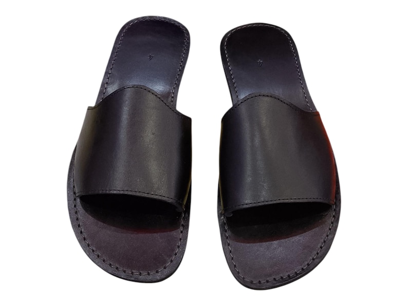 May include: A pair of black leather slide sandals. The sandals have a simple design with a wide strap across the top of the foot and a stitched sole. The sandals are a dark charcoal color.