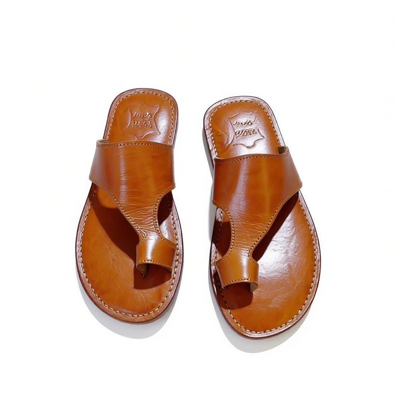May include: A pair of brown leather sandals with a toe loop design. The sandals have a smooth, polished finish and are stitched around the edges. The sandals are displayed on a white background.