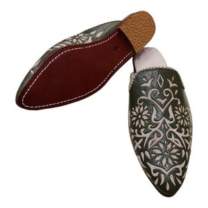 May include: A pair of green leather slippers with intricate floral and geometric cut-out designs. The slippers have a pointed toe, a brown sole, and a light pink interior. The stitching is white, and the overall style is traditional.