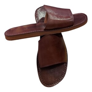 May include: A pair of brown leather sandals. The sandals have a simple design with a single strap across the top of the foot and a flat sole. The edges are stitched with white thread.