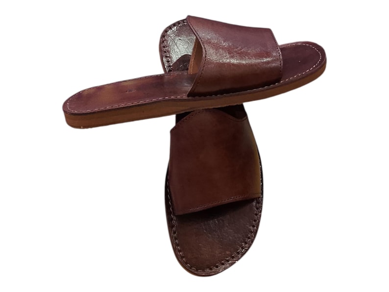 May include: A pair of brown leather sandals. The sandals have a simple design with a single strap across the top of the foot and a flat sole. The edges are stitched with white thread.