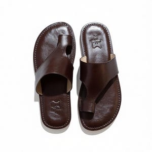 May include: A pair of brown leather sandals with a thong-style design. The sandals have a stitched border and a small embossed emblem near the heel. The sandals are designed for comfort and style.