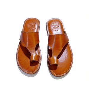 May include: A pair of brown leather sandals with a toe loop design. The sandals have a smooth, polished finish and are stitched around the edges. The sandals are displayed on a white background.