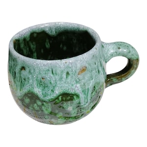 Może przedstawiać: Ręcznie wykonany ceramiczny kubek o zaokrąglonym kształcie i uchwycie. Kubek ma unikalne szkliwo w odcieniach zieleni i bieli, tworzące teksturowany, organiczny wygląd. Wnętrze jest ciemnozielone.