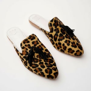 May include: A pair of leopard print slip-on mules. The shoes feature a tan and brown spotted pattern, black tassels, and white insoles. The soles are a light brown color.