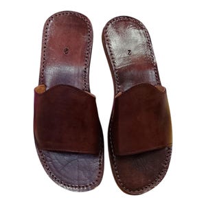 May include: A pair of brown leather sandals with a slip-on design. The sandals have a dark brown color and are stitched around the edges. The number "2" is visible on the top of each sandal. These sandals are suitable for casual wear.