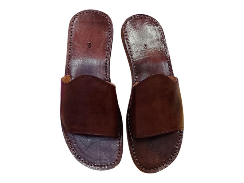 May include: A pair of brown leather sandals with a slip-on design. The sandals have a dark brown color and are stitched around the edges. The number "2" is visible on the top of each sandal. These sandals are suitable for casual wear.