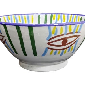 Può includere: Una ciotola in ceramica bianca con un bordo largo, con un design dipinto a mano. La ciotola presenta strisce verticali verdi e gialle, dettagli blu e motivi a occhio marrone. Il bordo è dipinto di viola.