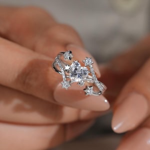 Peut inclure: Une bague argentée ornée d'une pierre centrale transparente en forme de cœur, entourée de plusieurs petits motifs en forme d'étoile. L'anneau présente un design croisé et est agrémenté de minuscules pierres pavées sur les côtés.