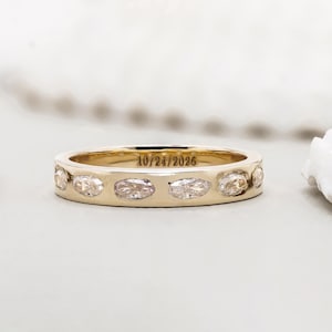 May include: A gold band ring featuring a series of horizontally set marquise-cut clear stones embedded along the outer surface. The interior of the band is engraved with the date 10/24/2026 in a clear, dark, sans-serif font style.