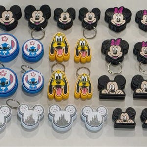 Puede incluir: Una colección de varios llaveros y portatarjetas con personajes como Mickey Mouse, Minnie Mouse, Pluto y Stitch. Los artículos incluyen diseños circulares, con formas y brillantes con anillas de metal, mostrando caras de dibujos animados.
