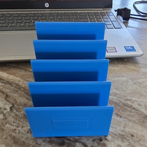 May include: A stack of four bright blue plastic desk organizers. Each organizer has a rectangular shape with a slightly angled front. The organizers are arranged in a stepped configuration.