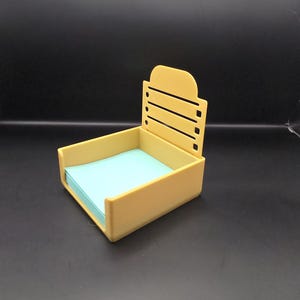May include: A light yellow desk organizer with a rectangular base and a back with horizontal slots. The organizer holds a stack of light blue notepaper. The design is simple and modern, suitable for office or home use.