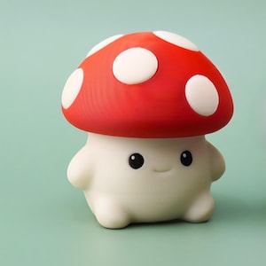 以下が含まれることがあります： 白いベースに赤い傘と白い水玉模様の愛らしいキノコの形をしたナイトライト。黒い目で笑顔の顔が特徴です。背景は明るい緑色です。