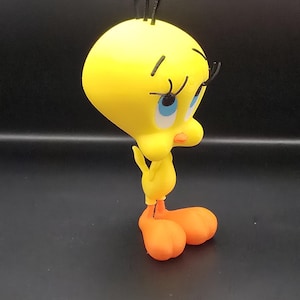 May include: A yellow Tweety Bird figurine with orange feet and a yellow body. The cartoon character has black hair and eyelashes, blue eyes, and an orange beak. The figurine is standing on a dark surface.