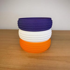 May include: Three stacked, textured headbands in vibrant colors: deep purple, crisp white, and bright orange. The headbands are made of a woven material, and are stacked on top of each other. The headbands are displayed on a wooden surface.
