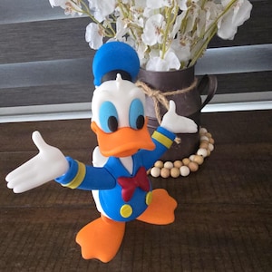 Könnte beinhalten: Eine Donald Duck-Figur in einem blauen Matrosenanzug, roter Fliege und orangefarbenen Füßen und Schnabel. Die Figur ist mit ausgestreckten Armen dargestellt. Im Hintergrund befindet sich eine Vase mit weißen Blumen.