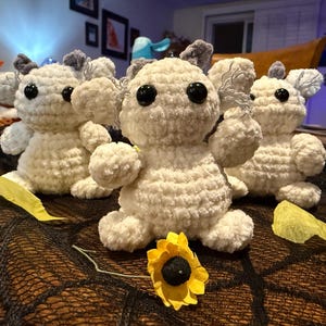 May include: Four handmade, off-white crochet stuffed animals with black button eyes and gray ears. They are arranged on a dark surface with a yellow sunflower and leaf accents. The toys have a soft, textured appearance.