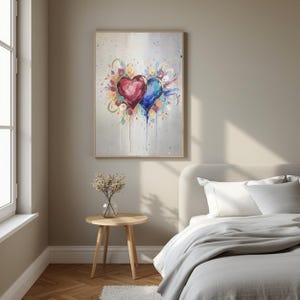 Puede incluir: Una obra de arte enmarcada con dos corazones de acuarela, uno rojo y otro azul, con acentos florales coloridos. La pintura tiene un fondo claro con salpicaduras de pintura y se exhibe en un dormitorio.