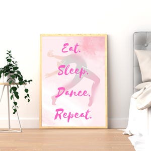 Könnte beinhalten: Ein gerahmter Druck mit den Worten "Eat. Sleep. Dance. Repeat." in rosa Schrift. Das Kunstwerk zeigt eine Tänzerin im Aquarellstil in Bewegung, mit einem weichen rosa und weißen Hintergrund. Der Rahmen ist hellholzfarben.