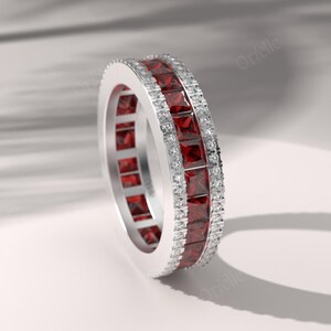 May include: A silver-toned eternity band featuring a central row of square-cut deep red gemstones flanked by two rows of small, clear, round-cut stones. The ring has a polished metallic finish and a continuous design that encircles the entire band.