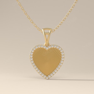 Op de afbeelding: Een goudkleurige hartvormige hangerketting. De hartvormige hanger heeft een gestructureerd midden met een zonnestraalontwerp en is omlijnd met kleine, heldere edelstenen. De ketting is ook goudkleurig.
