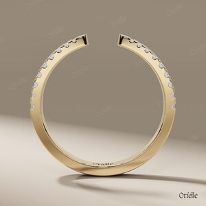 May include: A gold-colored ring with a unique open design. The ring is adorned with small, sparkling gemstones set along the outer curve. The brand name "Orelle" is subtly engraved on the ring's surface. The ring is displayed against a neutral background.