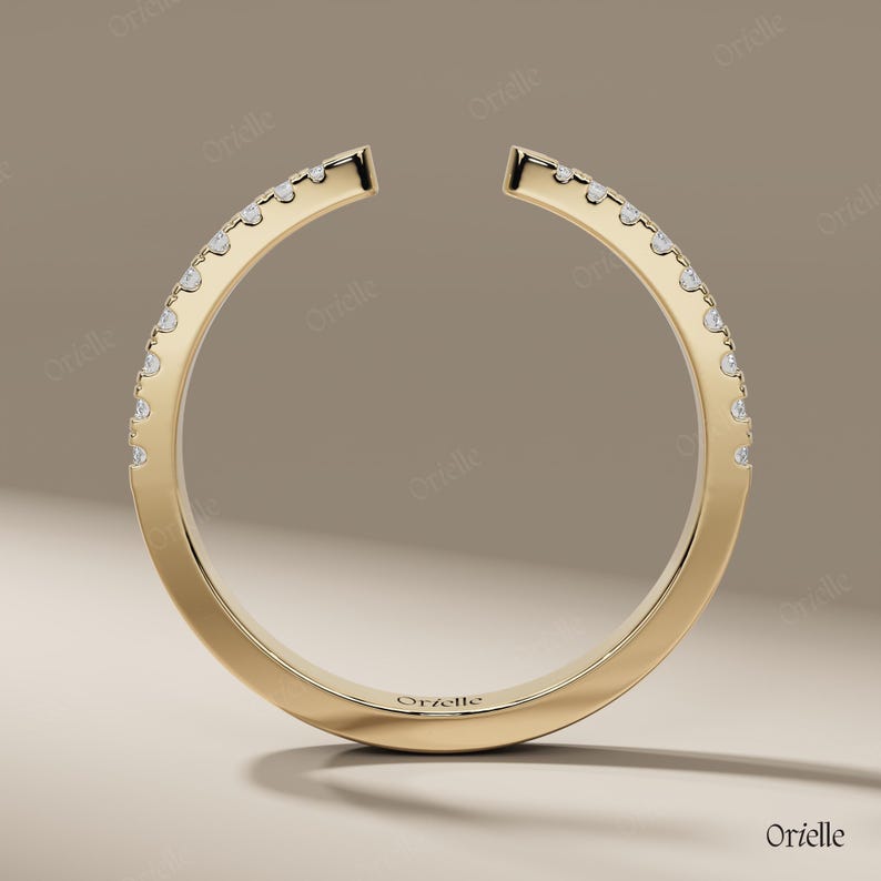 May include: A gold-colored ring with a unique open design. The ring is adorned with small, sparkling gemstones set along the outer curve. The brand name "Orelle" is subtly engraved on the ring's surface. The ring is displayed against a neutral background.