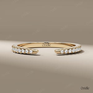 May include: A gold-colored ring with a row of small, clear gemstones set along the top half. The ring has an open design, with the ends not connected. The word "Orielle" is engraved on the inside of the ring.